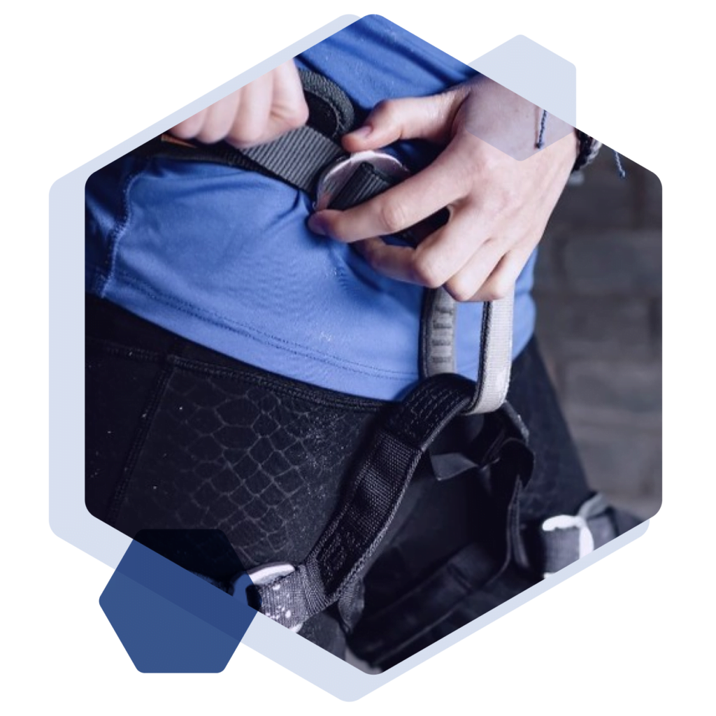 Eine Person passt einen Klettergurt an, während sie ein blaues Hemd trägt und vor einer grauen Ziegelwand steht.